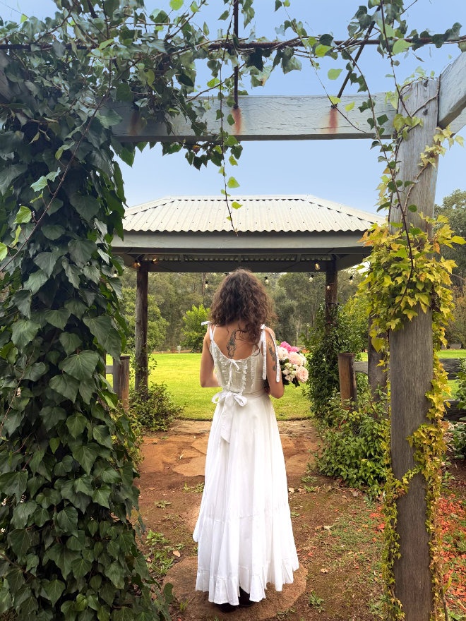 A bride in her wedding dress, holding a flower bouquet, stands in the Black Cockatoo Cellar Door garden, looking out to the lawn and forest trees.