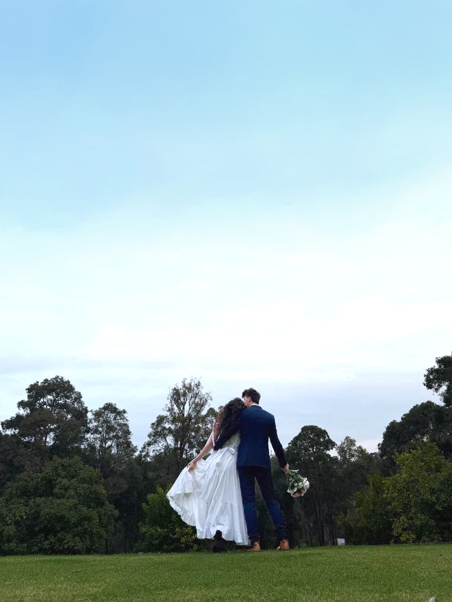 A wedding couple stands on the lawn at the Black Cockatoo Distillery Cellar Door, overlooking forest trees, under a big sky.