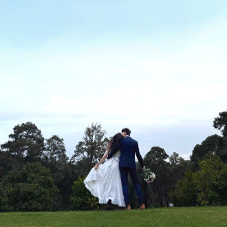 A wedding couple stands on the lawn at the Black Cockatoo Distillery Cellar Door, overlooking forest trees, under a big sky.