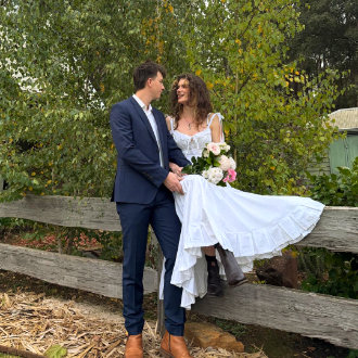 A wedding couple in the Black Cockatoo Distillery garden - the bride sits on the wooden fence holding hands with her groom standing close by.
